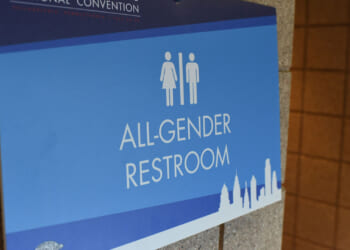 Blue sign on a wall reads All-Gender Restroom with icons of a woman and man. The bottom features a city skyline graphic.