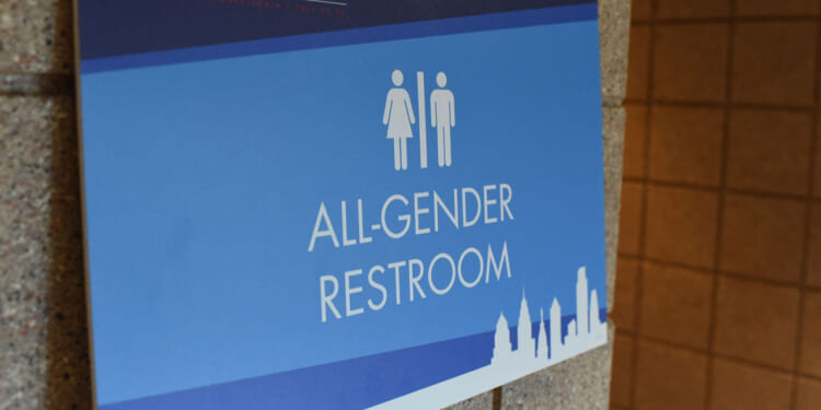 Blue sign on a wall reads All-Gender Restroom with icons of a woman and man. The bottom features a city skyline graphic.