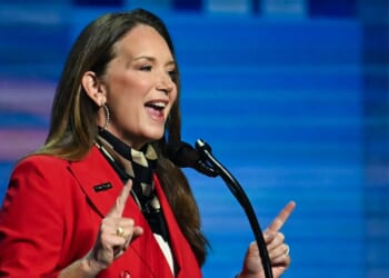 A woman in a red blazer speaks energetically at a microphone against a blue background, gesturing with both index fingers raised.
