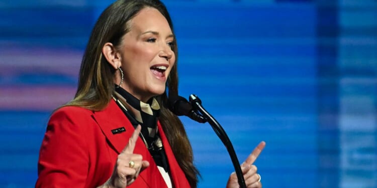 A woman in a red blazer speaks energetically at a microphone against a blue background, gesturing with both index fingers raised.