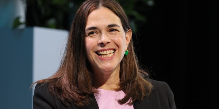 A woman with long brown hair, wearing a black blazer and pink top, smiles while seated indoors. Green plants are visible in the background.