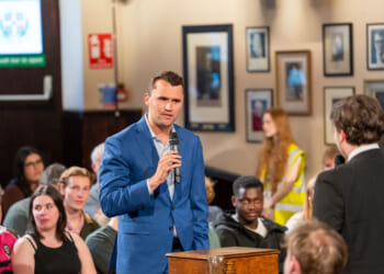A man in a blue suit speaks into a microphone during a discussion in a crowded room, with seated audience members and framed photos on the wall behind him.