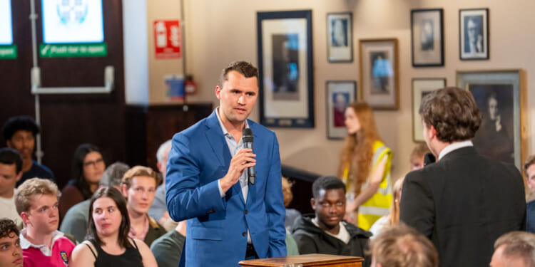 A man in a blue suit speaks into a microphone during a discussion in a crowded room, with seated audience members and framed photos on the wall behind him.