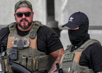 Two law enforcement officers in tactical gear stand together. One wears sunglasses and a cap, while the other wears a black mask and cap. Both have vests labeled with police insignia and various equipment attached.