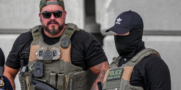 Two law enforcement officers in tactical gear stand together. One wears sunglasses and a cap, while the other wears a black mask and cap. Both have vests labeled with police insignia and various equipment attached.