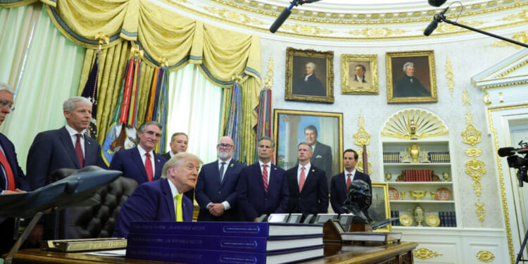 A group of men in suits, including Donald Trump seated at a desk, are gathered in the Oval Office with microphones overhead and shelves, flags, and presidential portraits in the background.