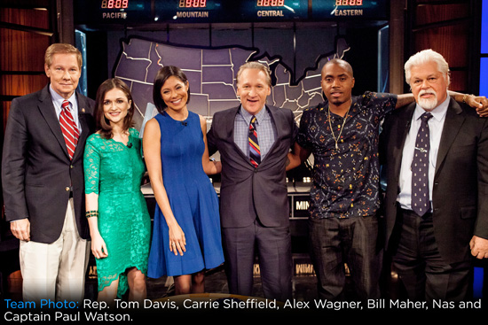 Six people stand together smiling in front of a U.S. time zone map on a TV studio set. They are dressed in business and semi-formal attire, with arms around each other, suggesting camaraderie and a group photo.