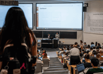 A large lecture hall filled with students facing two projection screens displaying a dashboard; instructors stand at the front. A periodic table hangs on the right wall. A student with a backpack stands in the foreground.