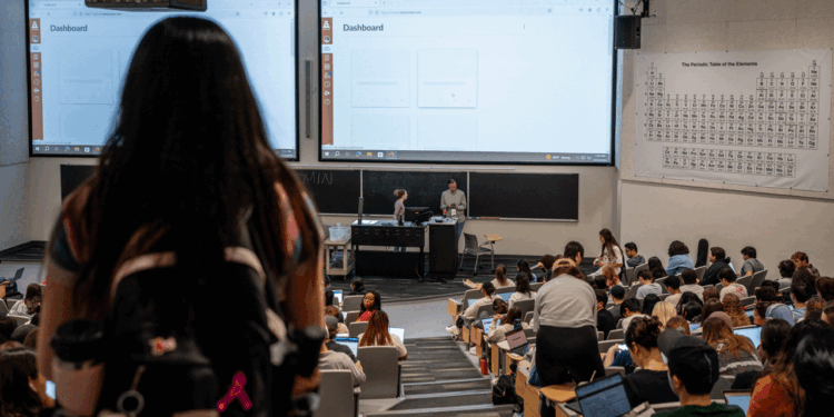 A large lecture hall filled with students facing two projection screens displaying a dashboard; instructors stand at the front. A periodic table hangs on the right wall. A student with a backpack stands in the foreground.