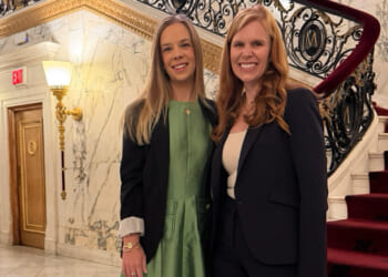 HHS officials Natalie Dodson and Bethany Kozma, one in a green dress and the other in a black suit, stand smiling in front of a grand marble staircase with ornate black railings and red carpet in an elegant building.