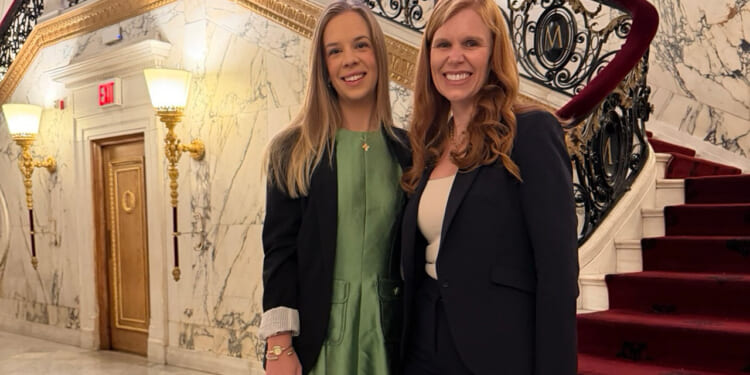 HHS officials Natalie Dodson and Bethany Kozma, one in a green dress and the other in a black suit, stand smiling in front of a grand marble staircase with ornate black railings and red carpet in an elegant building.