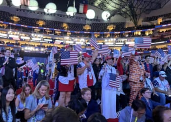 A crowded indoor arena at the Republican National Midterm Convention with people dressed in patriotic outfits, many holding small American flags.
