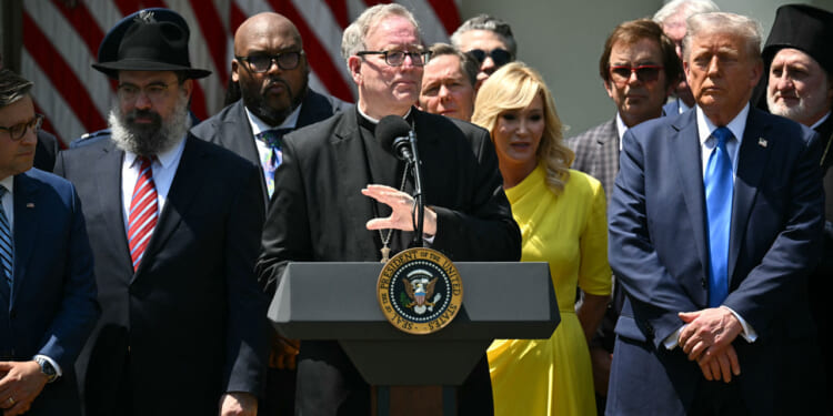 A group of people, including a man at a podium with the U.S. presidential seal, stand outdoors. American flags hang in the background. Some individuals wear suits, and one woman wears a bright yellow dress.