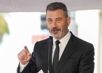 A man in a black suit and tie speaks into a microphone, gesturing with his right hand. Reminiscent of Kimmel’s charismatic stage presence, he has short, dark hair and a trimmed beard. The background is blurred, suggesting an outdoor event.