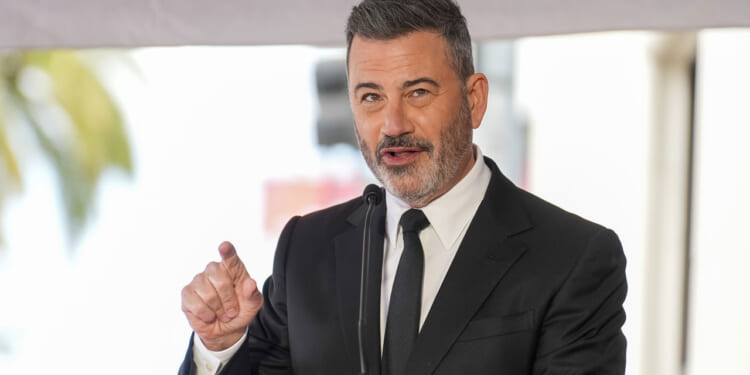 A man in a black suit and tie speaks into a microphone, gesturing with his right hand. Reminiscent of Kimmel’s charismatic stage presence, he has short, dark hair and a trimmed beard. The background is blurred, suggesting an outdoor event.