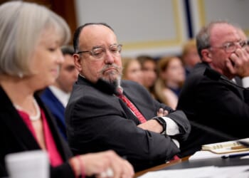 Three people sit at a hearing table, facing forward. The person in the center, wearing glasses and a suit, looks serious with arms crossed. The others appear to be listening intently, with papers and microphones in front of them.