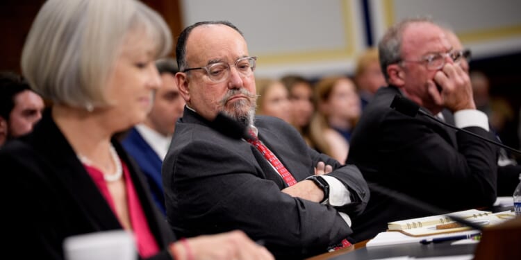Three people sit at a hearing table, facing forward. The person in the center, wearing glasses and a suit, looks serious with arms crossed. The others appear to be listening intently, with papers and microphones in front of them.