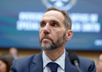 A man in a suit and tie with a beard looks to the side while seated in front of a blurred background with text.