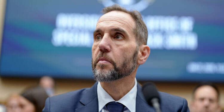 A man in a suit and tie with a beard looks to the side while seated in front of a blurred background with text.