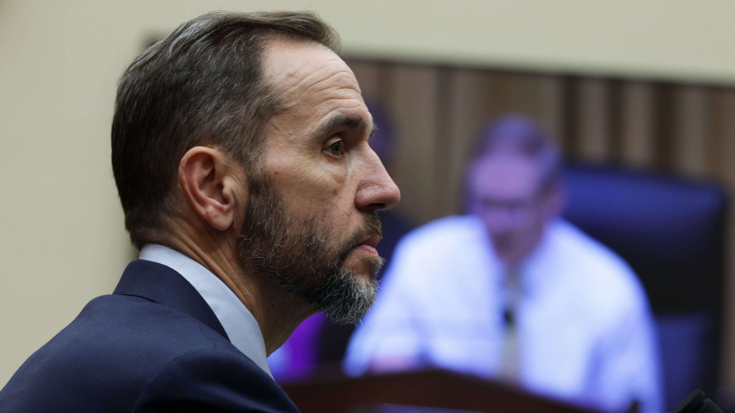 A man in a suit with a short beard listens intently during a meeting, while a blurry figure appears on a screen in the background.