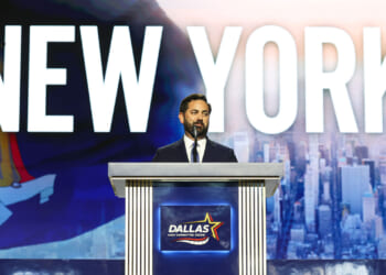 A man in a suit stands at a podium labeled Dallas with a cityscape and the word NEW YORK displayed on a large screen behind him.