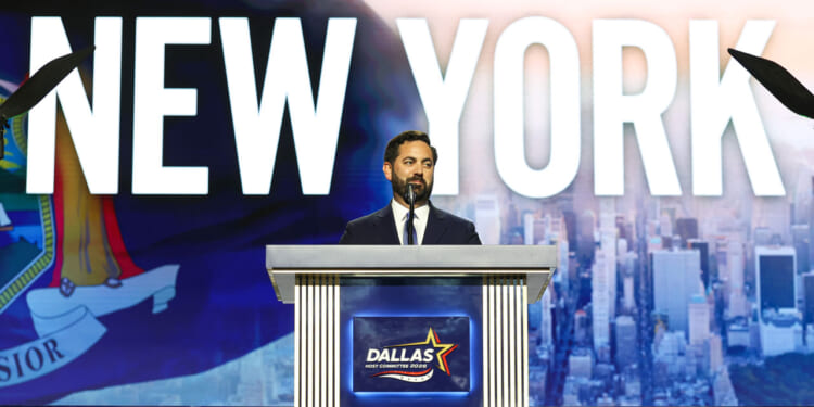 A man in a suit stands at a podium labeled Dallas with a cityscape and the word NEW YORK displayed on a large screen behind him.