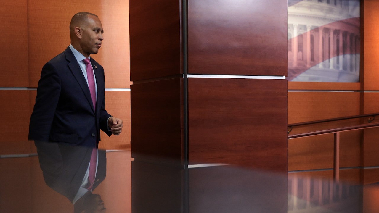 A man in a suit walks past a wooden wall inside a modern building, with a partial view of a white architectural structure reflected on the glossy surface below.