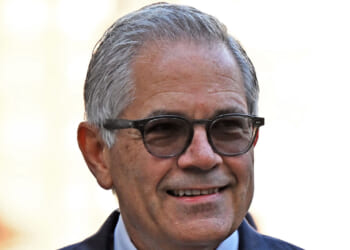 Smiling older man with gray hair and glasses wearing a suit, photographed outdoors with a blurred light background.