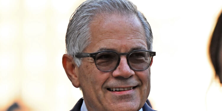 Smiling older man with gray hair and glasses wearing a suit, photographed outdoors with a blurred light background.