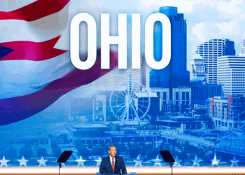 Jon Husted stands at a podium on stage with a large OHIO sign and cityscape backdrop, featuring a Ferris wheel and the Ohio flag.