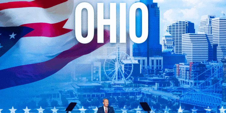 Jon Husted stands at a podium on stage with a large OHIO sign and cityscape backdrop, featuring a Ferris wheel and the Ohio flag.