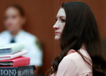 A woman with long brown hair sits in a courtroom, looking serious. Stacks of books and papers are in front of her, and a person in a white uniform is blurred in the background.