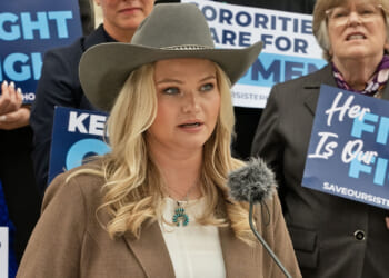 Kappa Kappa Gamma members, including Allie Coghan, sued KKG for allowing men to join the sorority. Pictured: Coghan, a woman in a cowboy hat speaks at a podium, surrounded by people holding blue signs with messages supporting women and sisterhood.