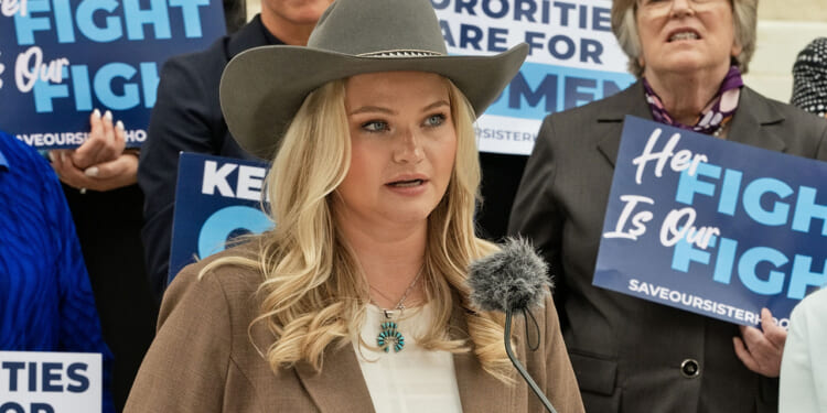 Kappa Kappa Gamma members, including Allie Coghan, sued KKG for allowing men to join the sorority. Pictured: Coghan, a woman in a cowboy hat speaks at a podium, surrounded by people holding blue signs with messages supporting women and sisterhood.