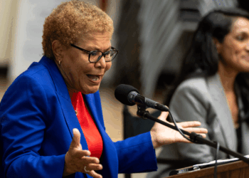 An older woman in a blue blazer speaks passionately into a microphone, gesturing with her hands. Another woman sits beside her, listening and smiling.