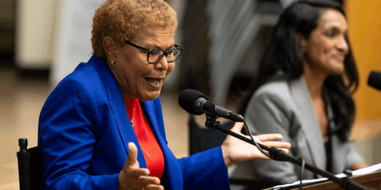 An older woman in a blue blazer speaks passionately into a microphone, gesturing with her hands. Another woman sits beside her, listening and smiling.