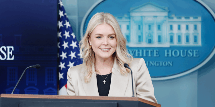 A woman with blond hair in a beige blazer speaks at a podium in the White House press briefing room, where past Trump administration officials often addressed the media. The American flag and the White House emblem are visible in the background.