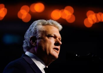 Rep. Mike Collins is pictured and speaks into a microphone, dressed in a suit, with warm, blurred orange lights in the background.