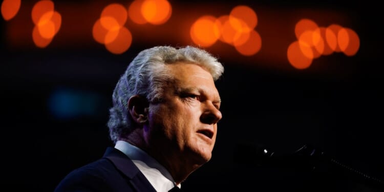 Rep. Mike Collins is pictured and speaks into a microphone, dressed in a suit, with warm, blurred orange lights in the background.