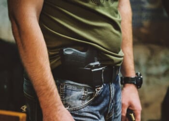 A person wearing jeans and a green shirt with a handgun holstered at their waist.