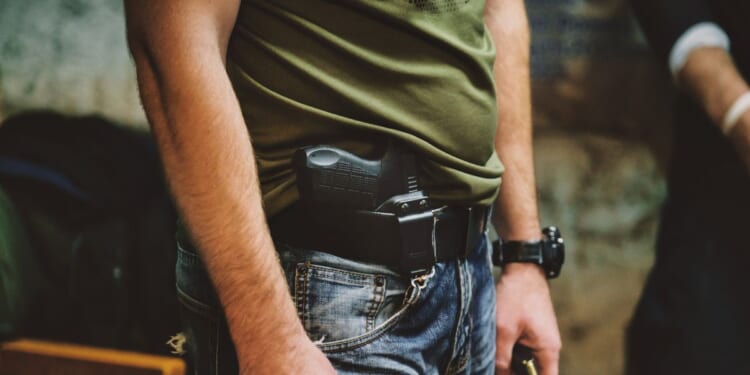 A person wearing jeans and a green shirt with a handgun holstered at their waist.