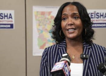 A woman in a striped blazer speaks at a podium with microphones. Behind her are campaign signs saying Keisha Lance Bottoms Governor and a colorful map on the wall.