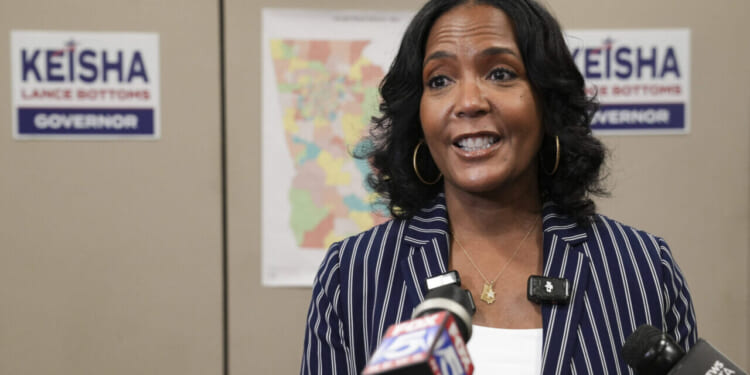 A woman in a striped blazer speaks at a podium with microphones. Behind her are campaign signs saying Keisha Lance Bottoms Governor and a colorful map on the wall.