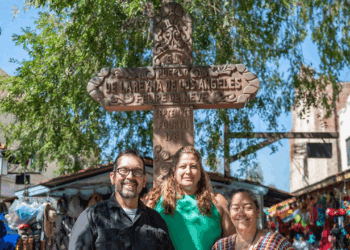 Three smiling people stand in front of a wooden cross monument in a sunny outdoor market with colorful goods and trees around them.