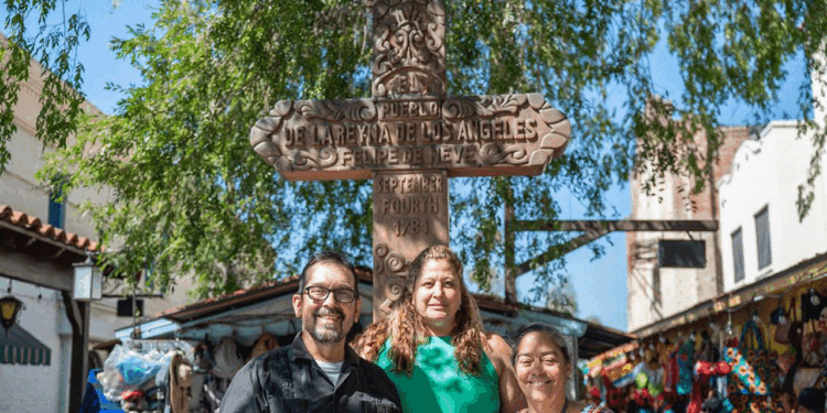 Three smiling people stand in front of a wooden cross monument in a sunny outdoor market with colorful goods and trees around them.