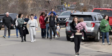 A group of adults and children walk together on a street near parked cars. One woman in front carries a child. Trees and emergency vehicles are in the background.