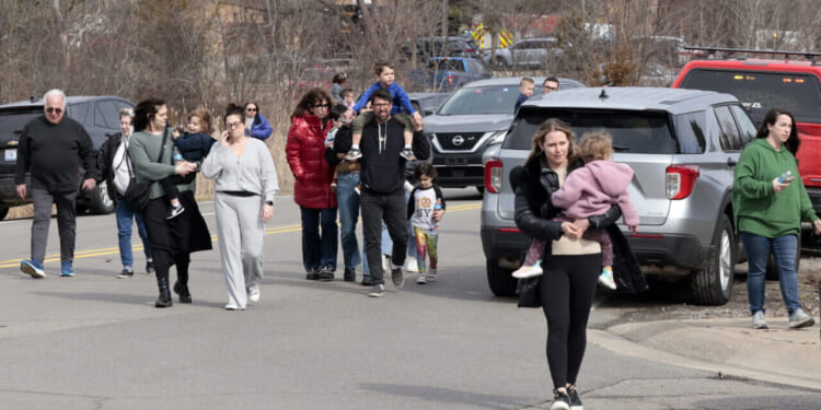 A group of adults and children walk together on a street near parked cars. One woman in front carries a child. Trees and emergency vehicles are in the background.