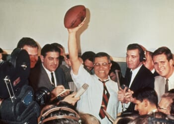 A smiling man in a tie triumphantly holds a football above his head, surrounded by reporters, cameras, and microphones in a crowded room.