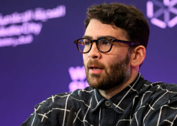 A man with dark hair and glasses, wearing a black and white checkered shirt, speaks on stage with a purple background featuring white geometric logos and Arabic text.