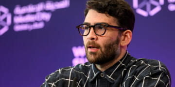 A man with dark hair and glasses, wearing a black and white checkered shirt, speaks on stage with a purple background featuring white geometric logos and Arabic text.